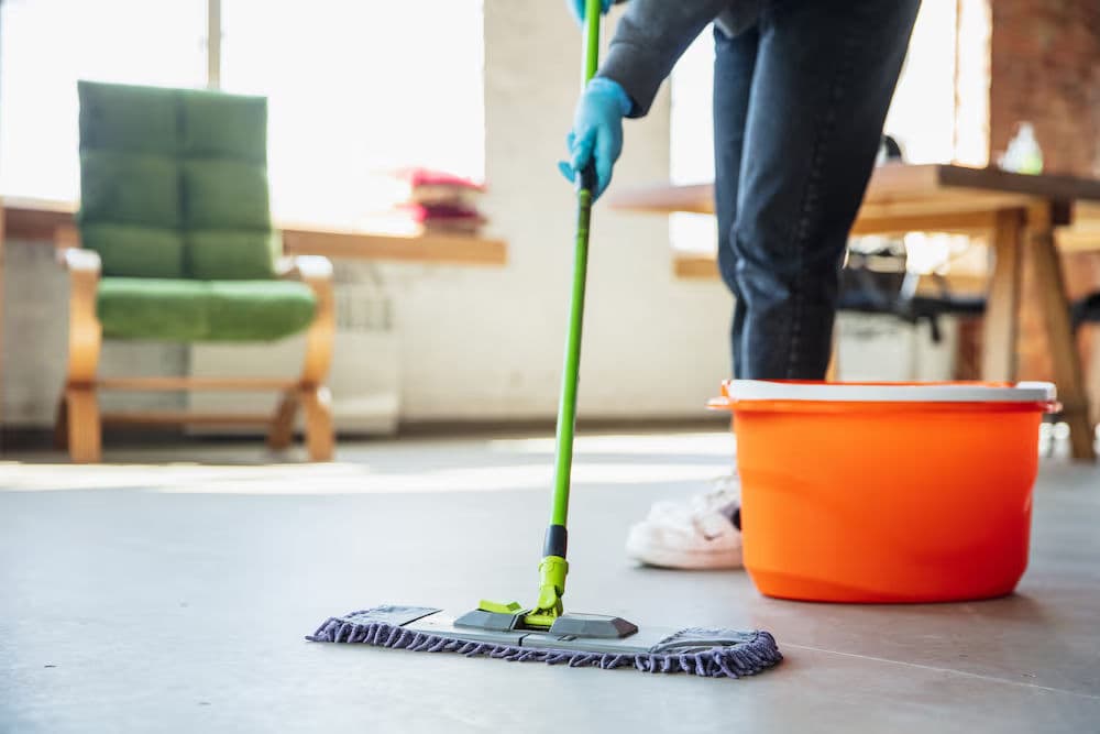 Person using a mop on a clean floor with an orange bucket in a bright indoor space.